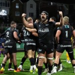 Ospreys players in black celebrating a try on the St Helen’s pitch at night, one with both fists raised