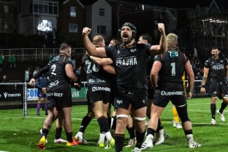 Ospreys players in black celebrating a try on the St Helen’s pitch at night, one with both fists raised