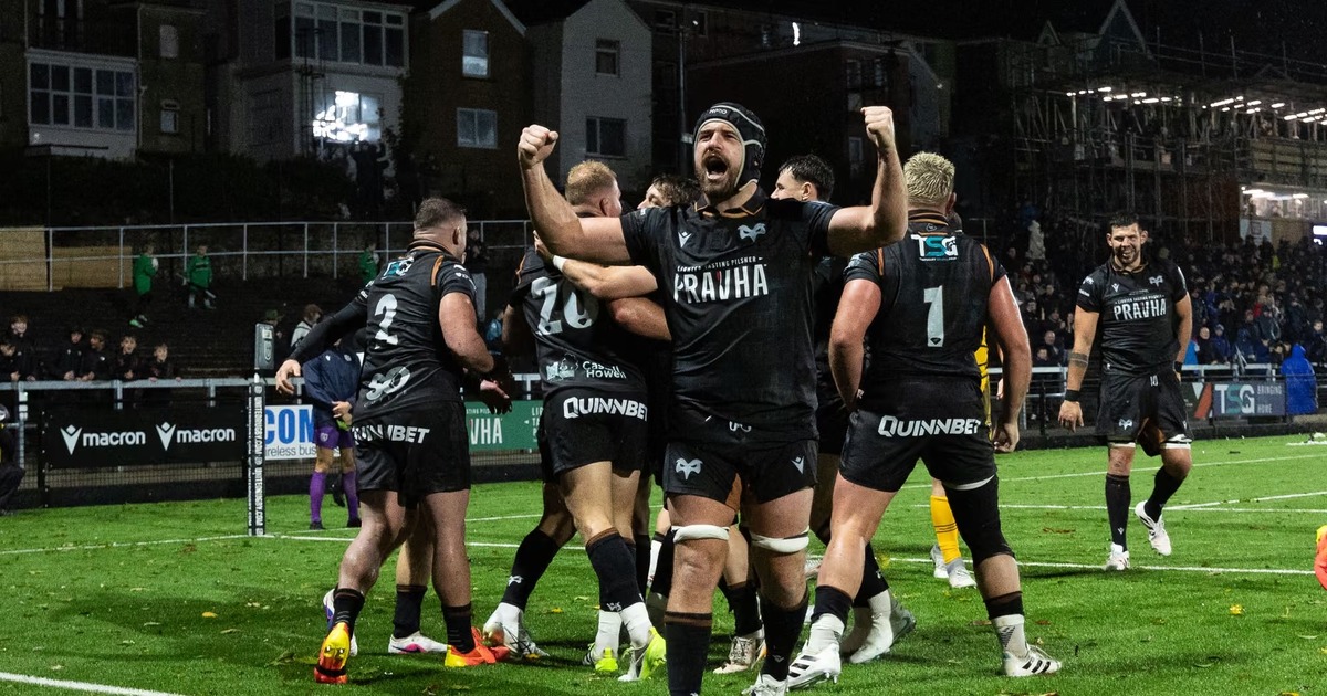 Ospreys players in black celebrating a try on the St Helen’s pitch at night, one with both fists raised