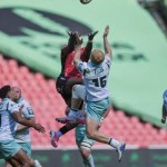 An Ospreys full-back in a white and teal kit leaps for a high ball against a Lions player in red at Ellis Park, with players from both sides watching