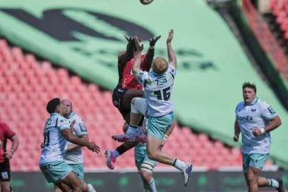 An Ospreys full-back in a white and teal kit leaps for a high ball against a Lions player in red at Ellis Park, with players from both sides watching