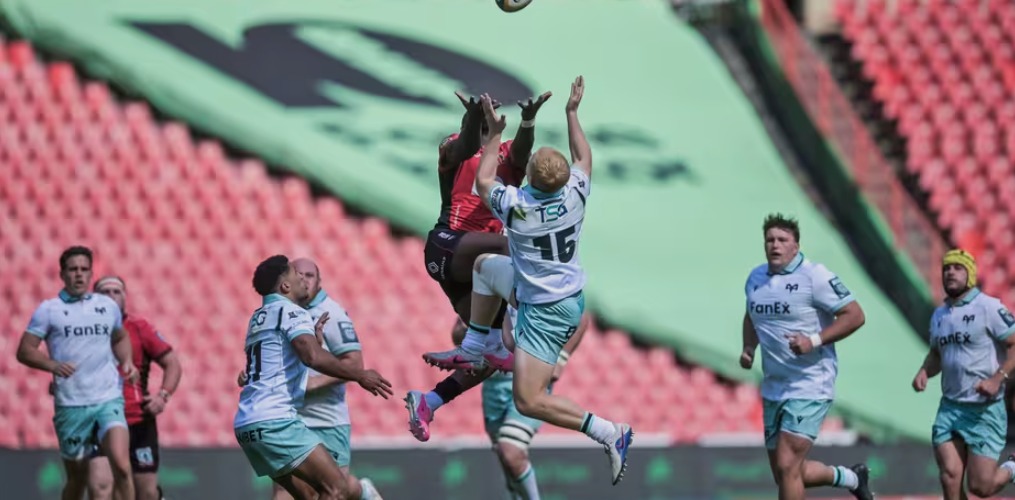 An Ospreys full-back in a white and teal kit leaps for a high ball against a Lions player in red at Ellis Park, with players from both sides watching