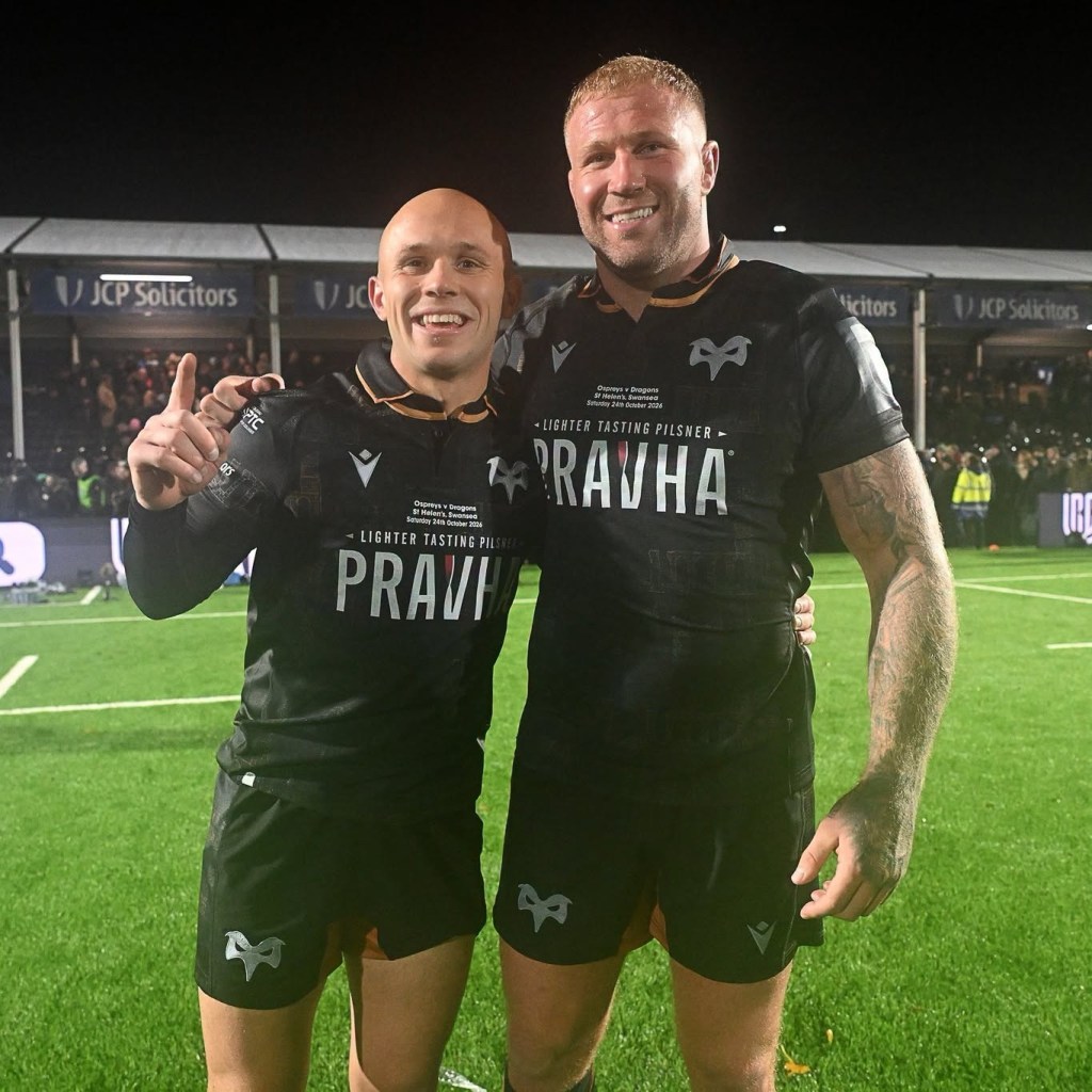 Two Ospreys players smiling for the camera on the pitch, one pointing a finger in the air