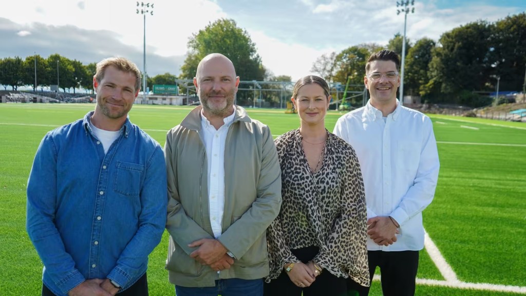 Four people standing on the new artificial pitch at St Helen's with the floodlights and stand behind them