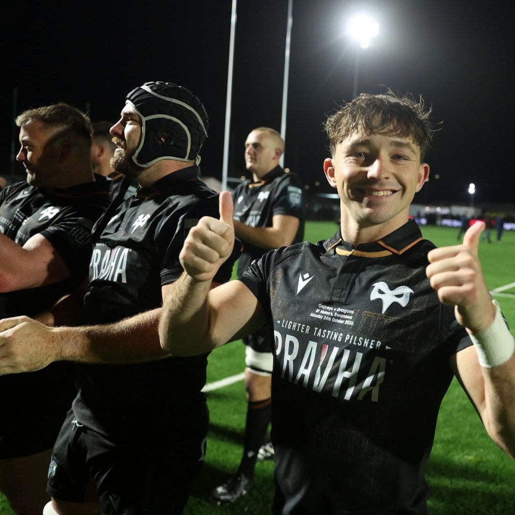 A smiling Ospreys player giving two thumbs up to the camera beside his team-mates