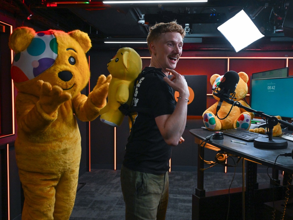 Owain Wyn Evans in the Radio 2 studio wearing the yellow Pudsey backpack, laughing, with a Pudsey mascot waving beside him and a Pudsey bear on the desk