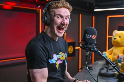 Owain Wyn Evans laughing in headphones at a BBC Radio 2 microphone, wearing a black Children in Need T-shirt, with a large Pudsey bear behind him