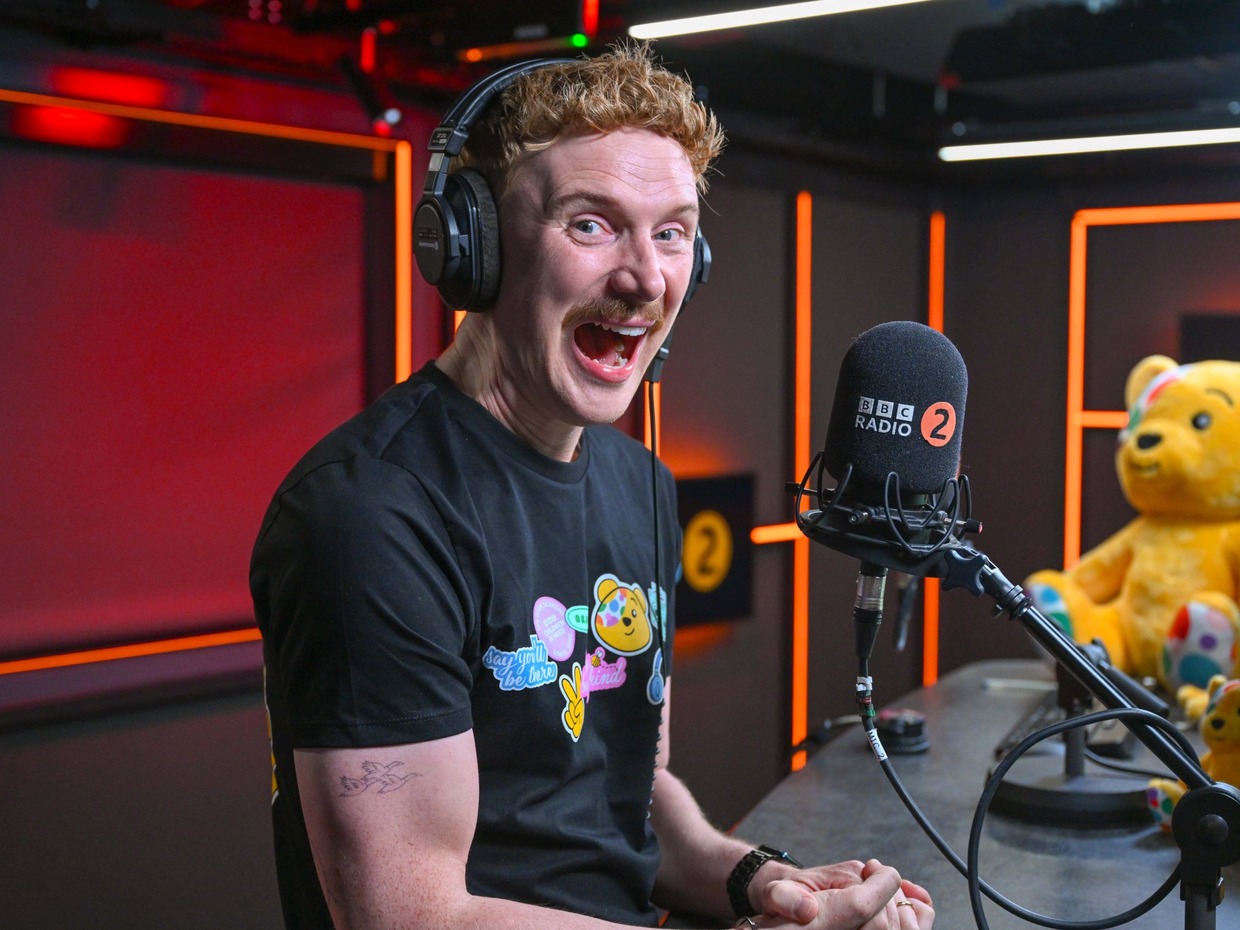 Owain Wyn Evans laughing in headphones at a BBC Radio 2 microphone, wearing a black Children in Need T-shirt, with a large Pudsey bear behind him