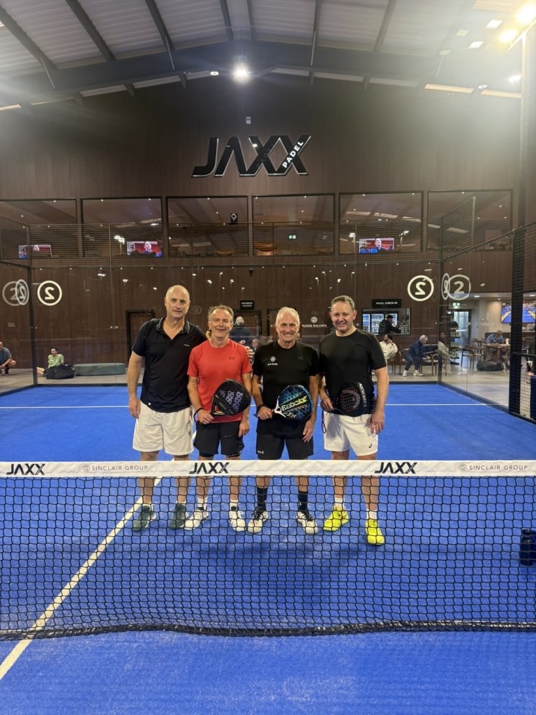 Four men holding padel rackets at the net on a blue court under the Jaxx Padel sign at the UK Padel Masters Series in Llansamlet