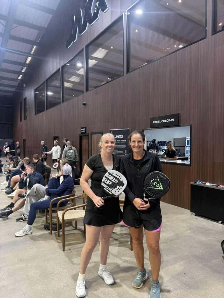 Two women holding padel rackets in front of the padel check-in desk at Jaxx Padel in Llansamlet, with spectators seated behind them