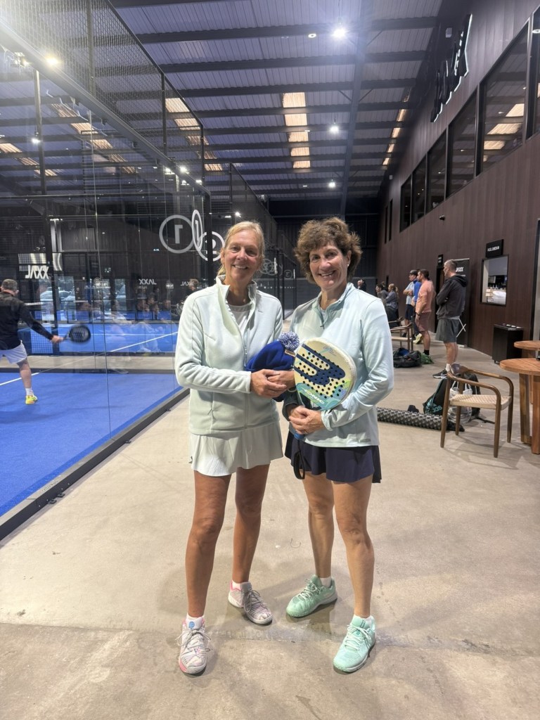 Two women players in pale blue tops holding a padel racket and a bobble hat beside the glass of a court at Jaxx Padel in Llansamlet