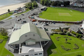 Aerial view of the Patti Pavilion with its curved green roof in Victoria Park, Swansea, with the St Helen's ground, Mumbles Road and the beach behind