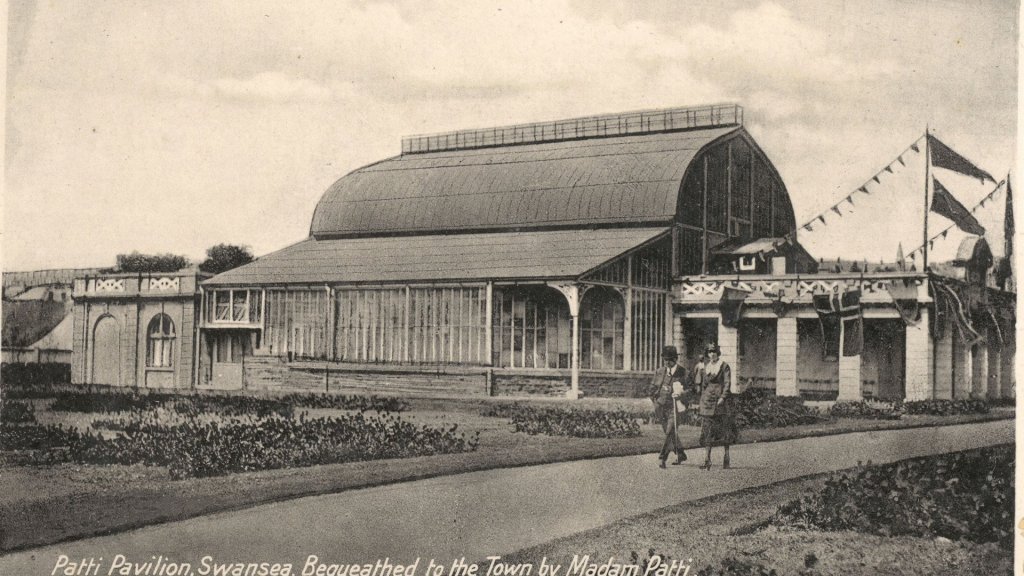 Old black and white postcard of the Patti Pavilion with a curved roof and glass walls, and a couple walking past on the path