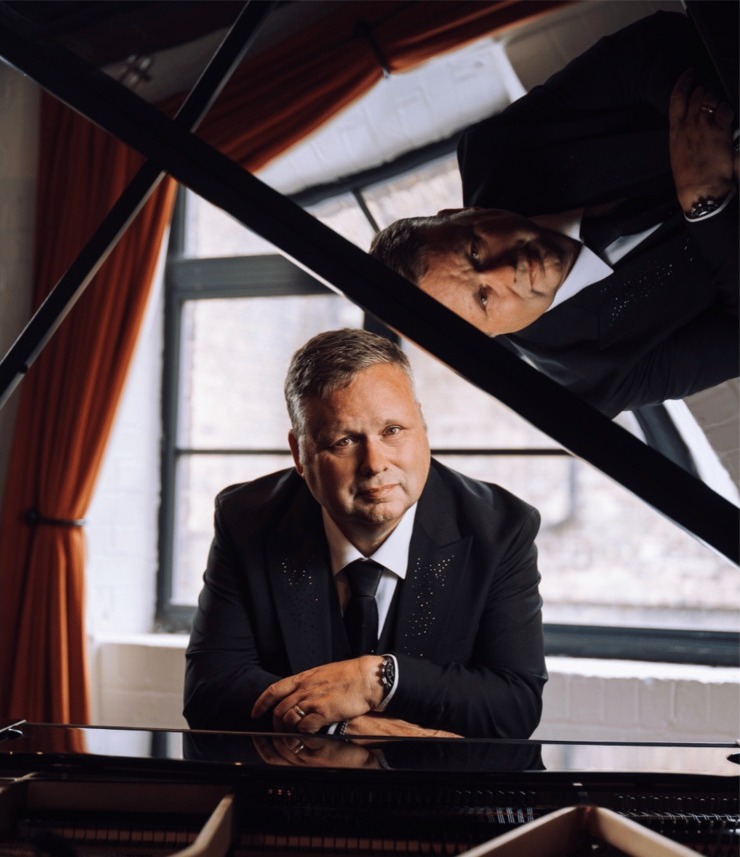 Paul Potts in a dark suit leaning on a grand piano, his reflection in the raised lid