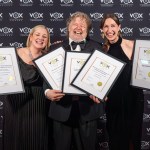 Nikki Mills, Phil Rowe and Jess Nesling laugh on the red carpet holding five framed VOX Awards certificates