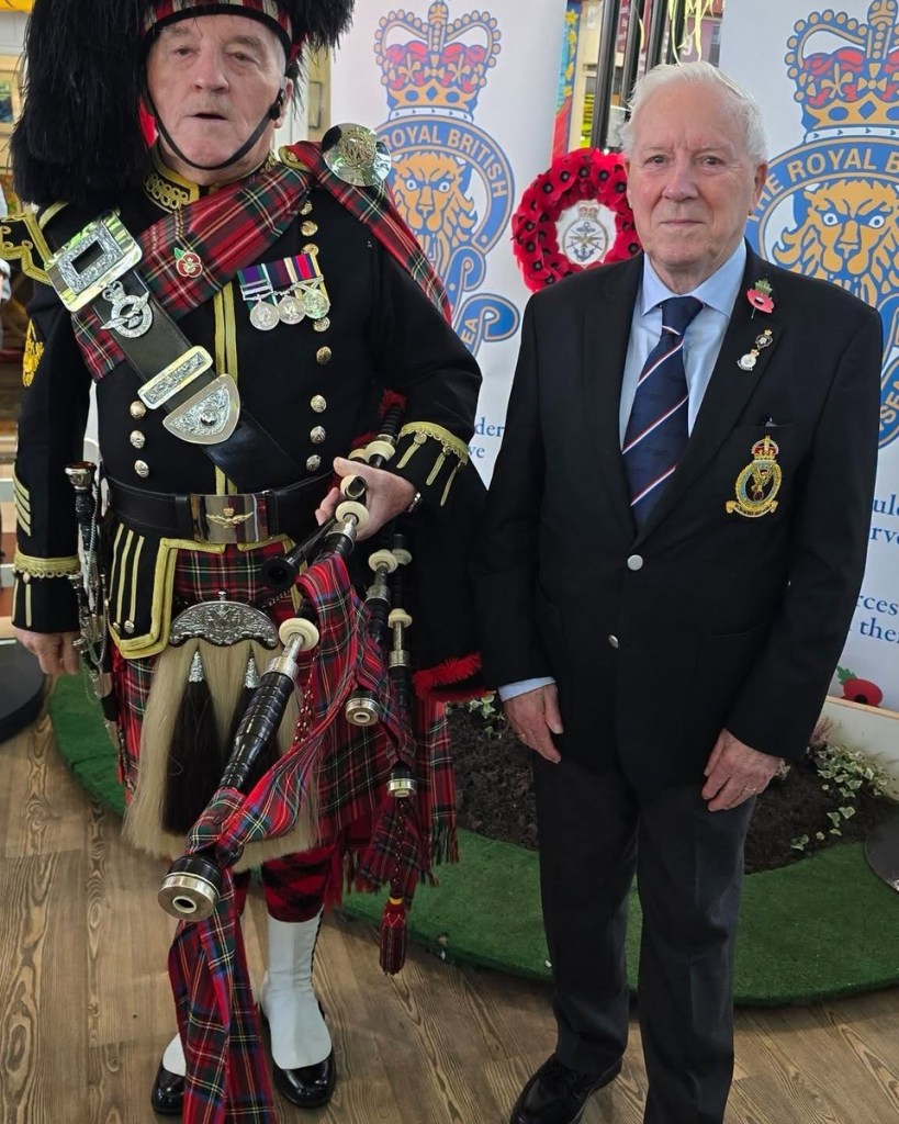 A piper in full Highland dress with bagpipes and medals stands beside a veteran in a blazer and poppy in front of Royal British Legion banners.