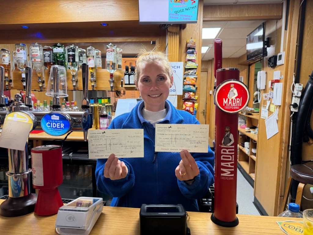 A woman behind the bar at Pontarddulais Conservative Club holding up two £250 cheques from the quiz charity account