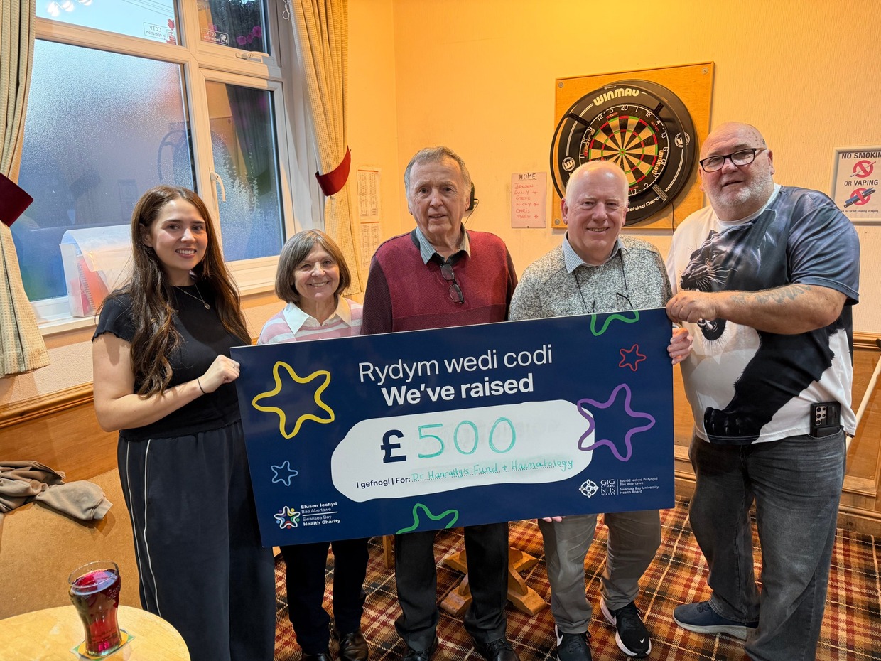 Five people holding a large Swansea Bay Health Charity cheque for £500 in front of a dartboard at Pontarddulais Conservative Club