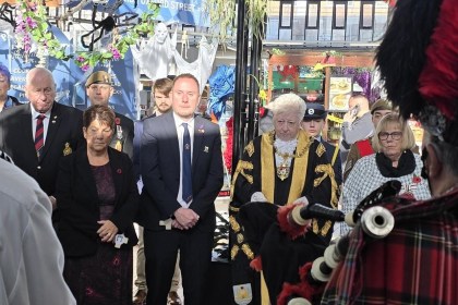 Council leader Rob Stewart and the Lord Mayor of Swansea in black and gold robes stand with veterans and guests in Swansea Market as a piper plays in the foreground.