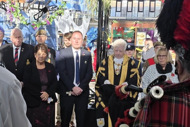 Council leader Rob Stewart and the Lord Mayor of Swansea in black and gold robes stand with veterans and guests in Swansea Market as a piper plays in the foreground.