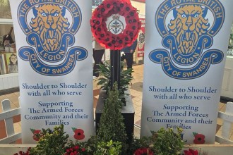 Two Royal British Legion City of Swansea banners either side of a poppy wreath, above a planted bed of cyclamen and heather in Swansea Market.