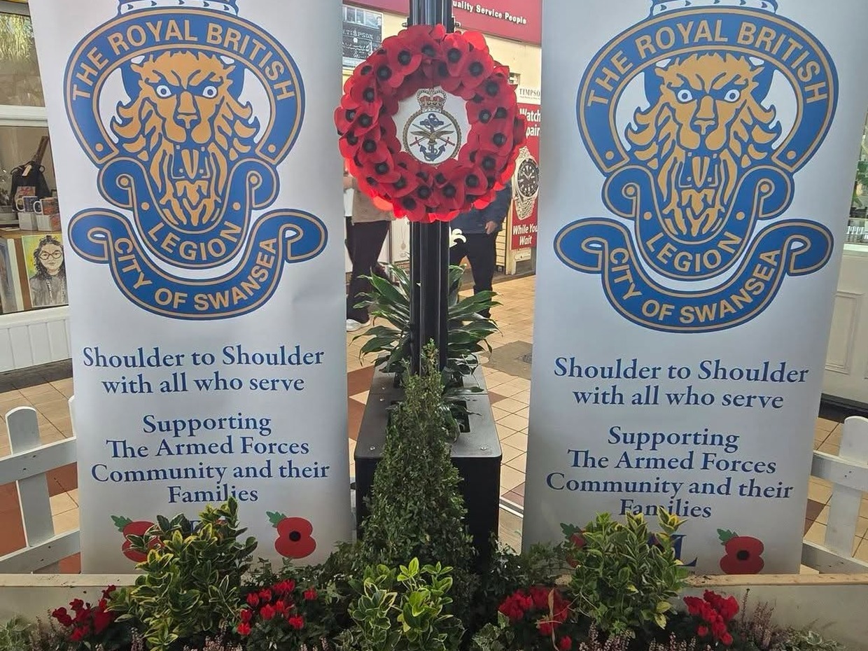 Two Royal British Legion City of Swansea banners either side of a poppy wreath, above a planted bed of cyclamen and heather in Swansea Market.