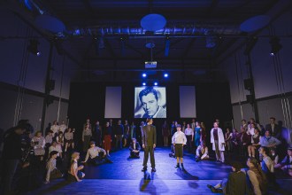 A large community cast in period costume on a blue-lit stage, with a black-and-white portrait of Richard Burton projected on the screen behind them