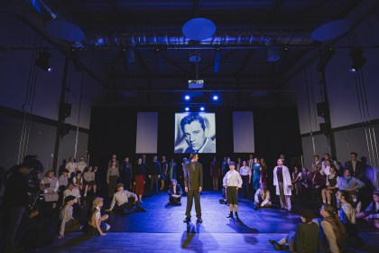 A large community cast in period costume on a blue-lit stage, with a black-and-white portrait of Richard Burton projected on the screen behind them