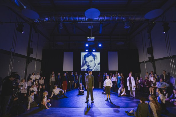 A large community cast in period costume on a blue-lit stage, with a black-and-white portrait of Richard Burton projected on the screen behind them