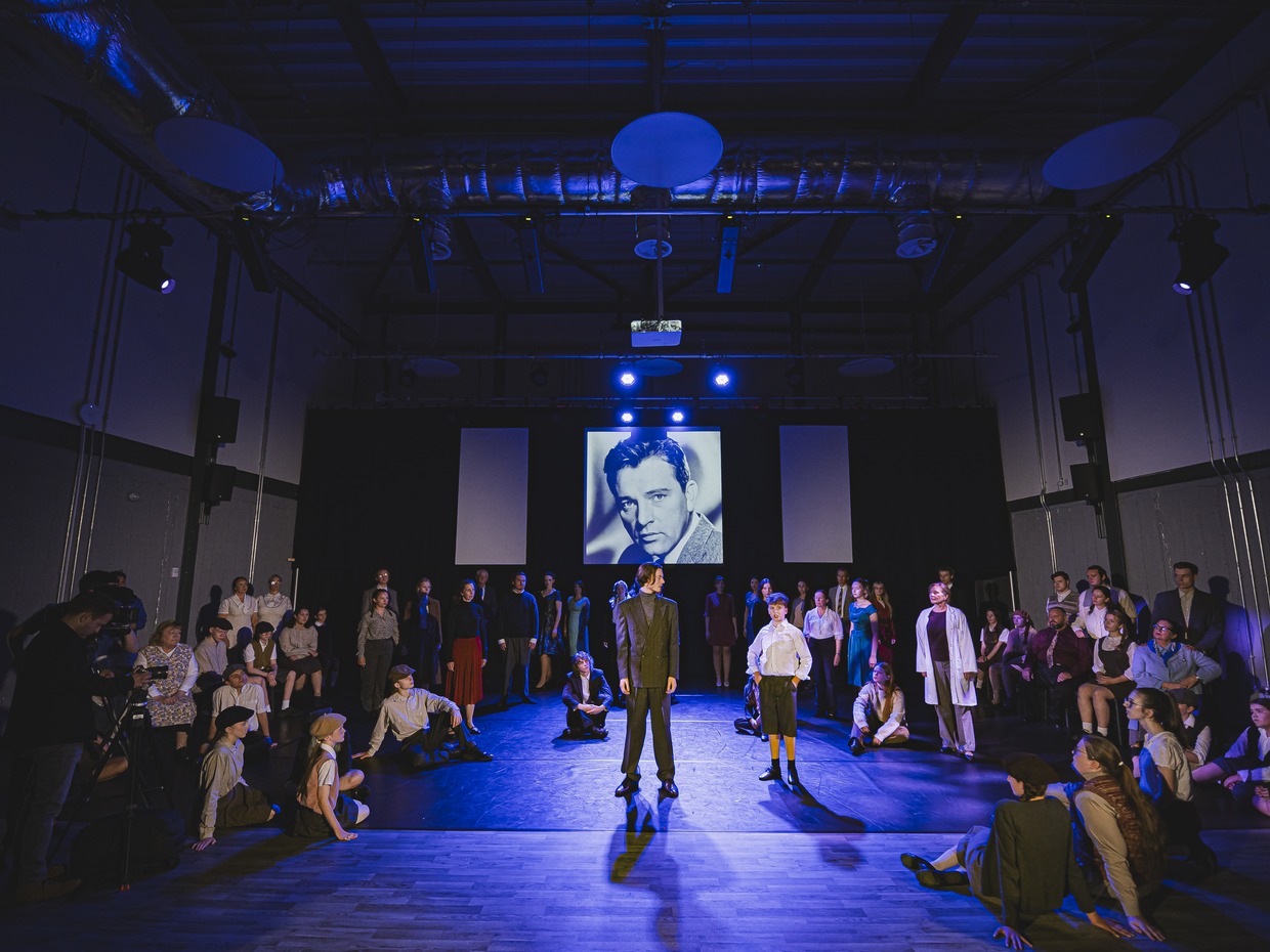 A large community cast in period costume on a blue-lit stage, with a black-and-white portrait of Richard Burton projected on the screen behind them