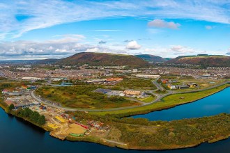 Aerial view of the Port Talbot waterfront at Harbourside, with industrial yards and scrubland beside the water and the town and hills behind.