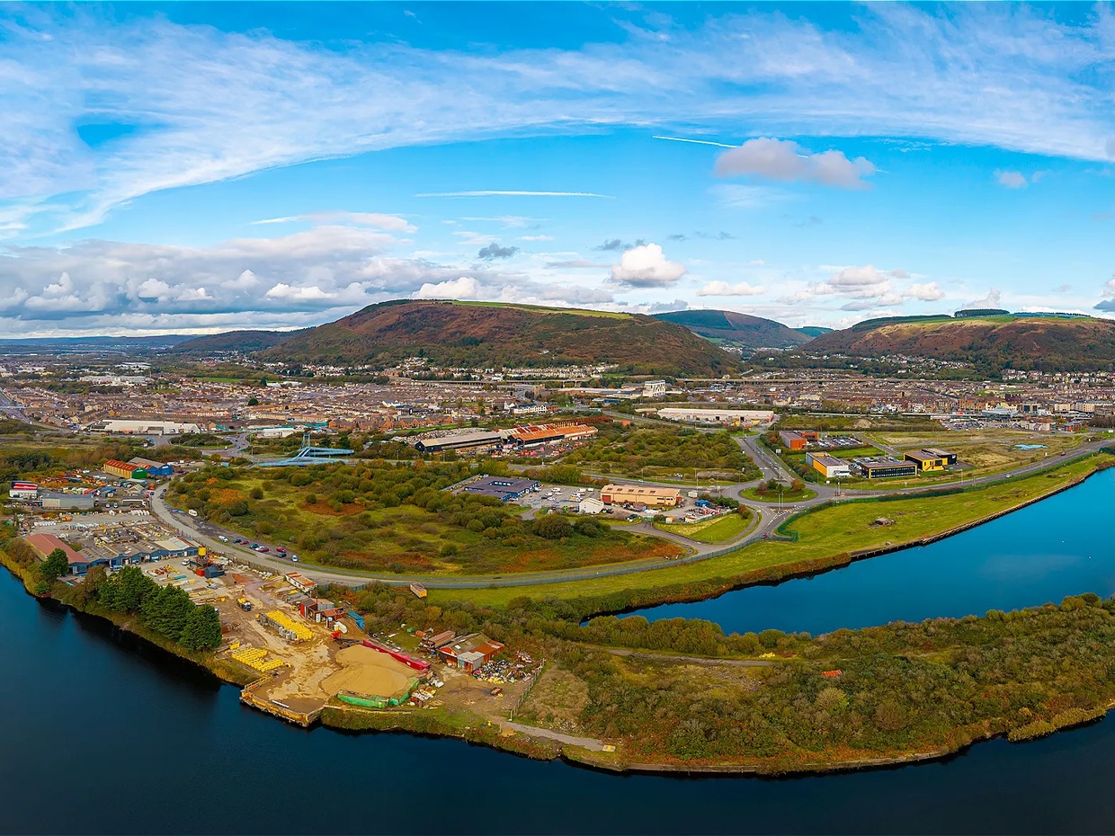 Aerial view of the Port Talbot waterfront at Harbourside, with industrial yards and scrubland beside the water and the town and hills behind.