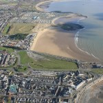 Aerial view of Porthcawl seafront, with the town in the foreground, the harbour on the right and a wide sandy beach curving away towards caravans in the distance