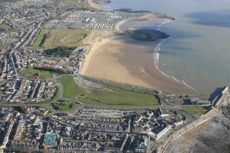 Aerial view of Porthcawl seafront, with the town in the foreground, the harbour on the right and a wide sandy beach curving away towards caravans in the distance