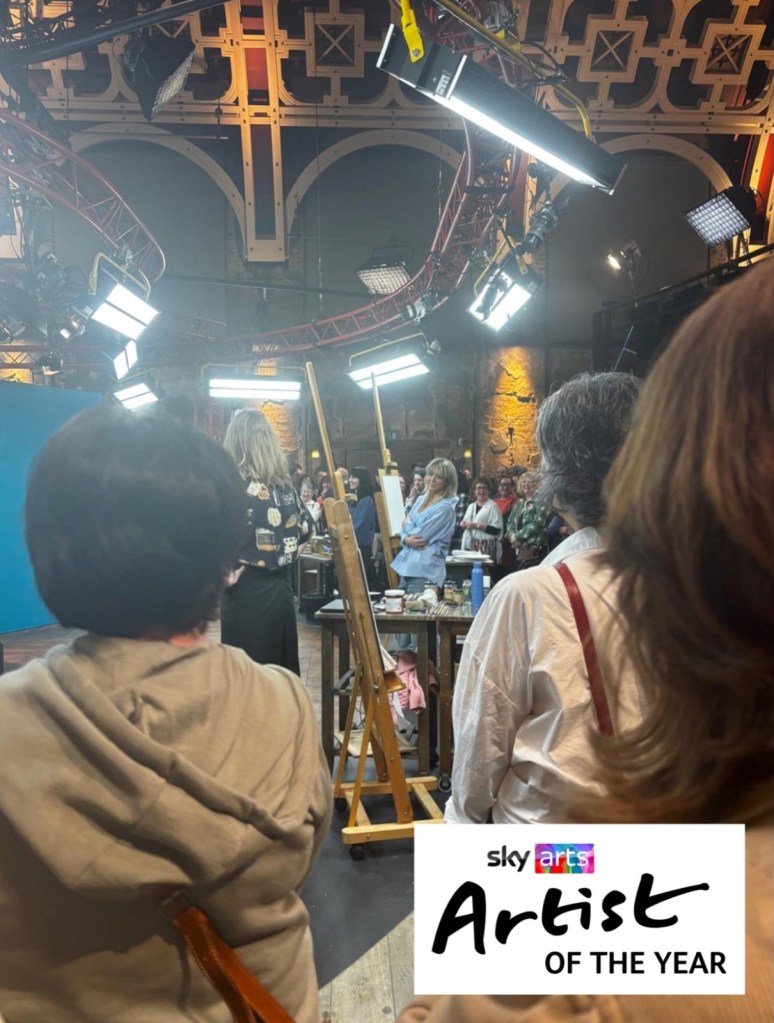 View over the heads of audience members towards easels and artists on a TV set under bright lights and an arched, patterned ceiling