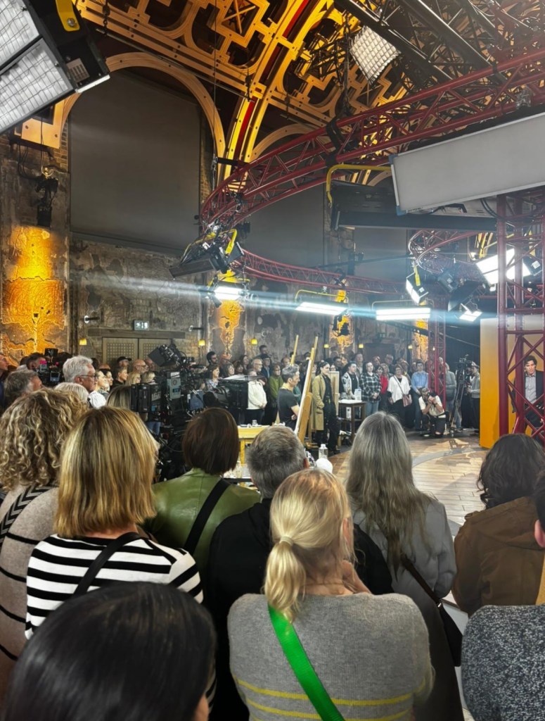 Rows of people seen from behind, packed around a TV set with easels, cameras and bright studio lights hanging from a red rig