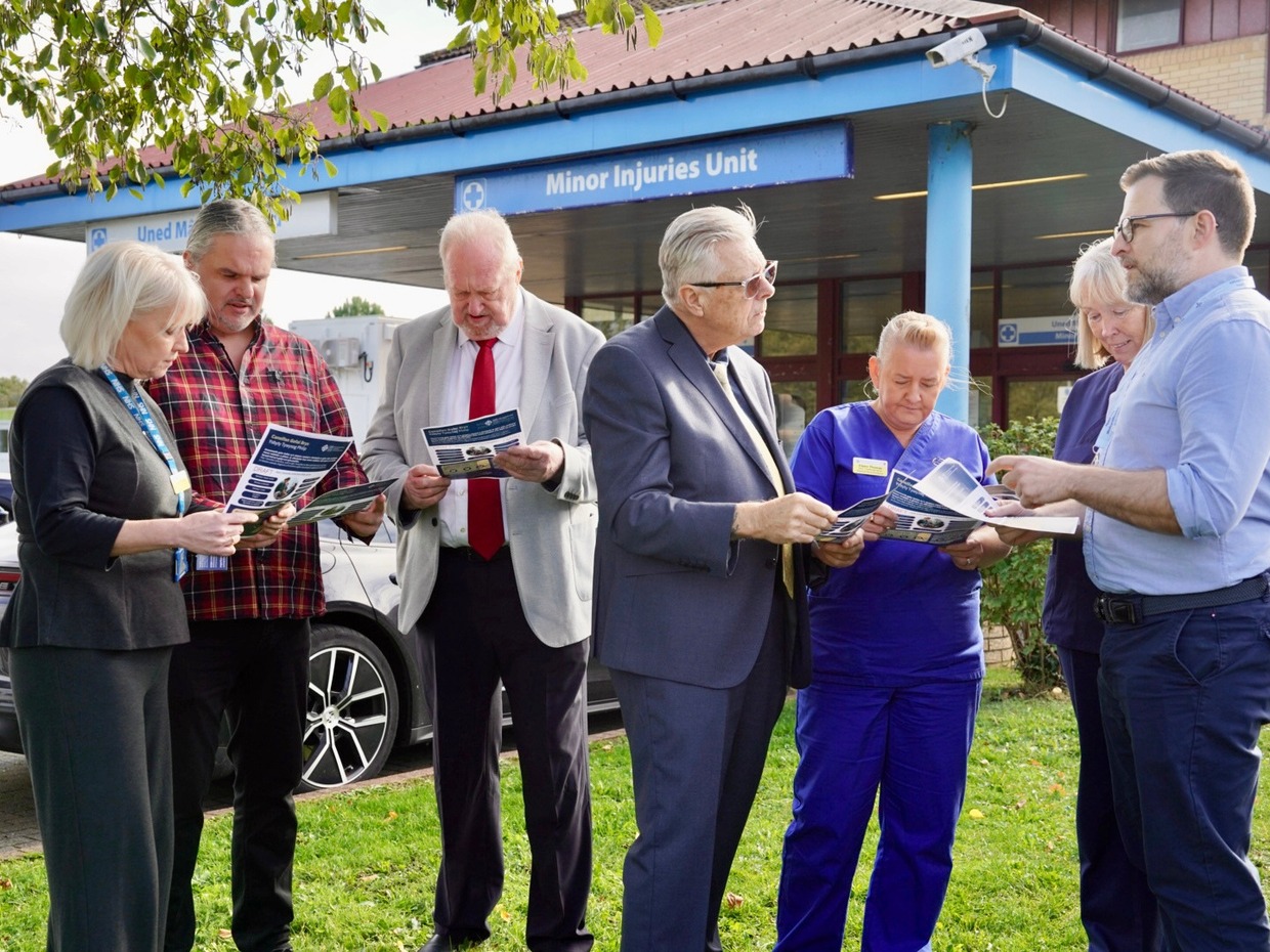 Seven people reading draft leaflets outside the Minor Injuries Unit entrance at Prince Philip Hospital in Llanelli, including SOSPPAN campaigners, a Llais representative and nursing staff in blue uniforms