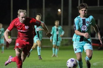Afan Lido's Alex Giardelli in red and Pure Swansea's Harvey Chapman in the teal away kit chasing the ball under the floodlights