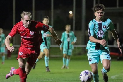 Afan Lido's Alex Giardelli in red and Pure Swansea's Harvey Chapman in the teal away kit chasing the ball under the floodlights
