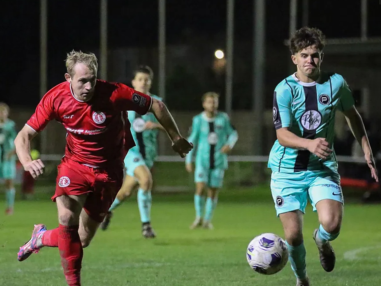 Afan Lido's Alex Giardelli in red and Pure Swansea's Harvey Chapman in the teal away kit chasing the ball under the floodlights
