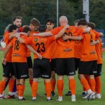 Pure Swansea players in orange shirts gathered in a huddle on the pitch at Swansea Bay Sports Park