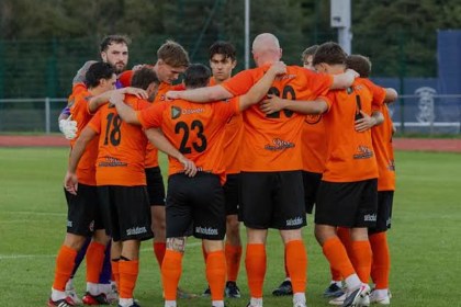 Pure Swansea players in orange shirts gathered in a huddle on the pitch at Swansea Bay Sports Park