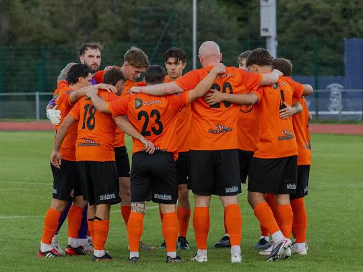 Pure Swansea players in orange shirts gathered in a huddle on the pitch at Swansea Bay Sports Park