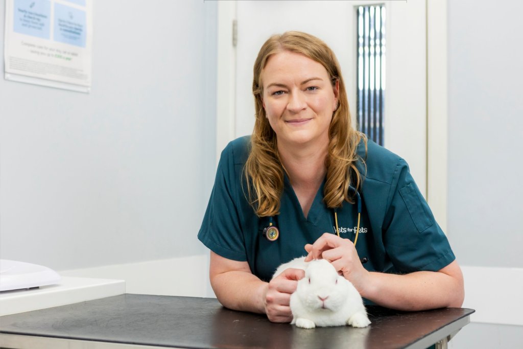 Vet Rebecca Madge in green scrubs with a stethoscope, leaning on an examination table and stroking a white rabbit