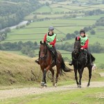 Two riders in red jackets and green bibs ride a bay and a black horse up a hillside track above a river valley in mid Wales