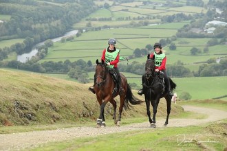 Two riders in red jackets and green bibs ride a bay and a black horse up a hillside track above a river valley in mid Wales