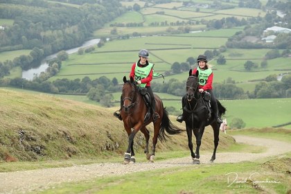 Two riders in red jackets and green bibs ride a bay and a black horse up a hillside track above a river valley in mid Wales