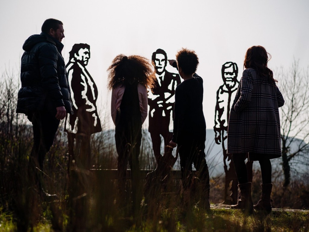 A family of four silhouetted at dusk standing among rusted steel cut-out figures of men on the Richard Burton trail, hills behind