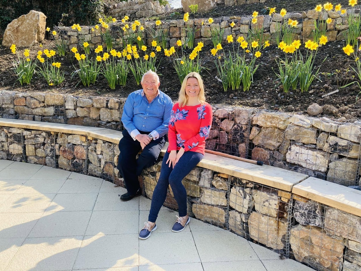 Cllr Richard Lewis and his daughter Francesca O'Brien sitting on a stone-filled gabion wall in front of a bank of daffodils