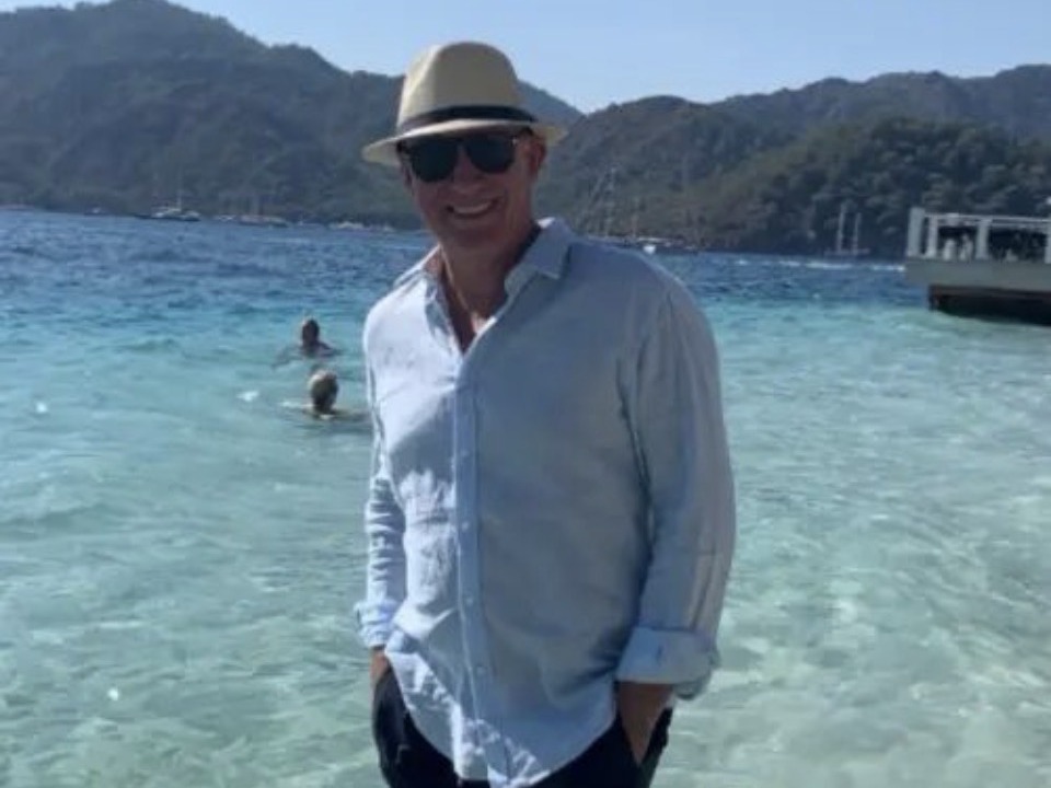 Rico Maza in a straw hat, sunglasses and a pale blue linen shirt, standing in clear shallow water